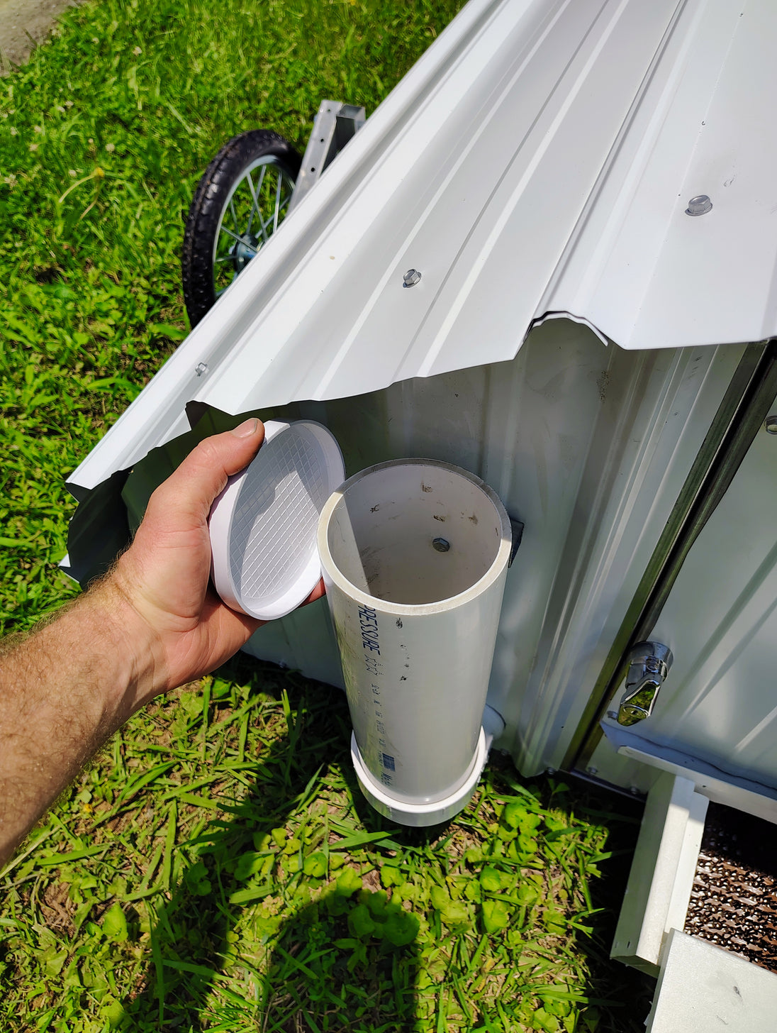 Person holding a lid to a white container for chicken feeder on a portable metal chicken coop on grass